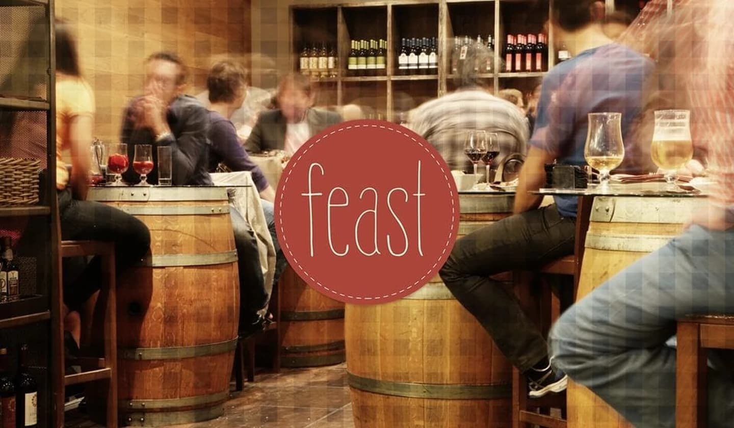 Feast wine bar identity over a warmly lit interior of patrons at wine-barrel tables.
