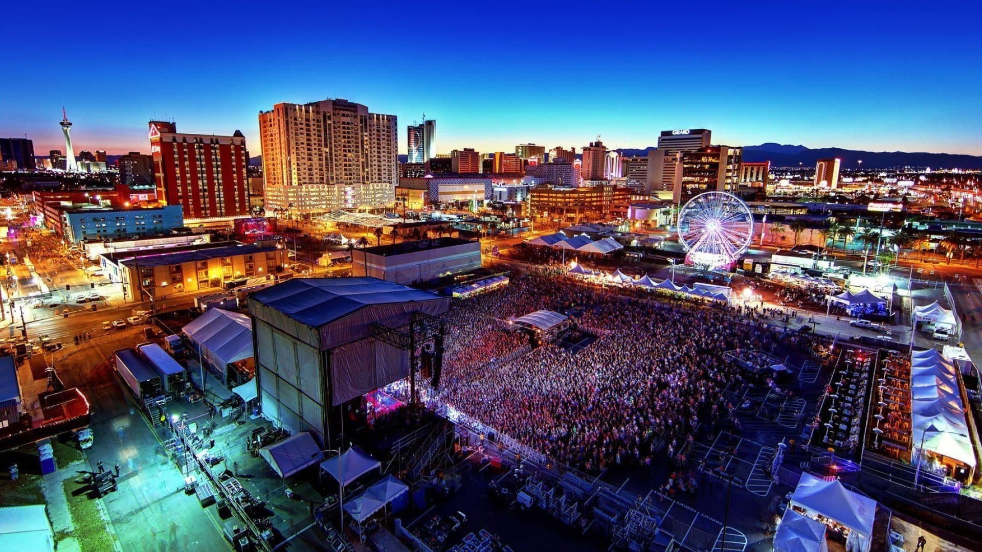 Aerial twilight photograph of the Life is Beautiful festival in downtown Las Vegas, with a packed main-stage crowd, a lit ferris wheel, and the city skyline beyond.