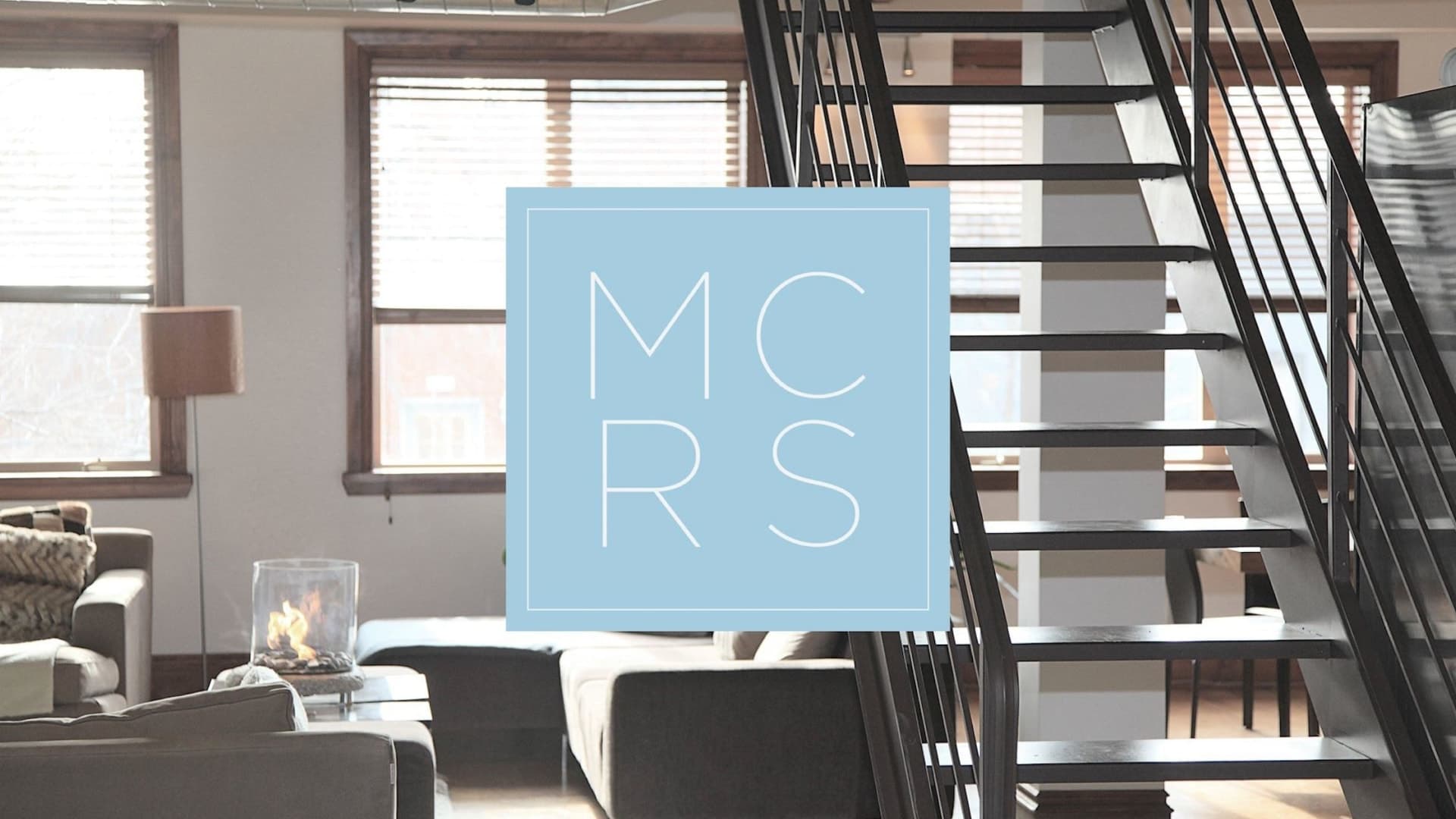 MCRS brand mark in pale blue set inside a square frame over a sunlit loft interior with an open black metal staircase.