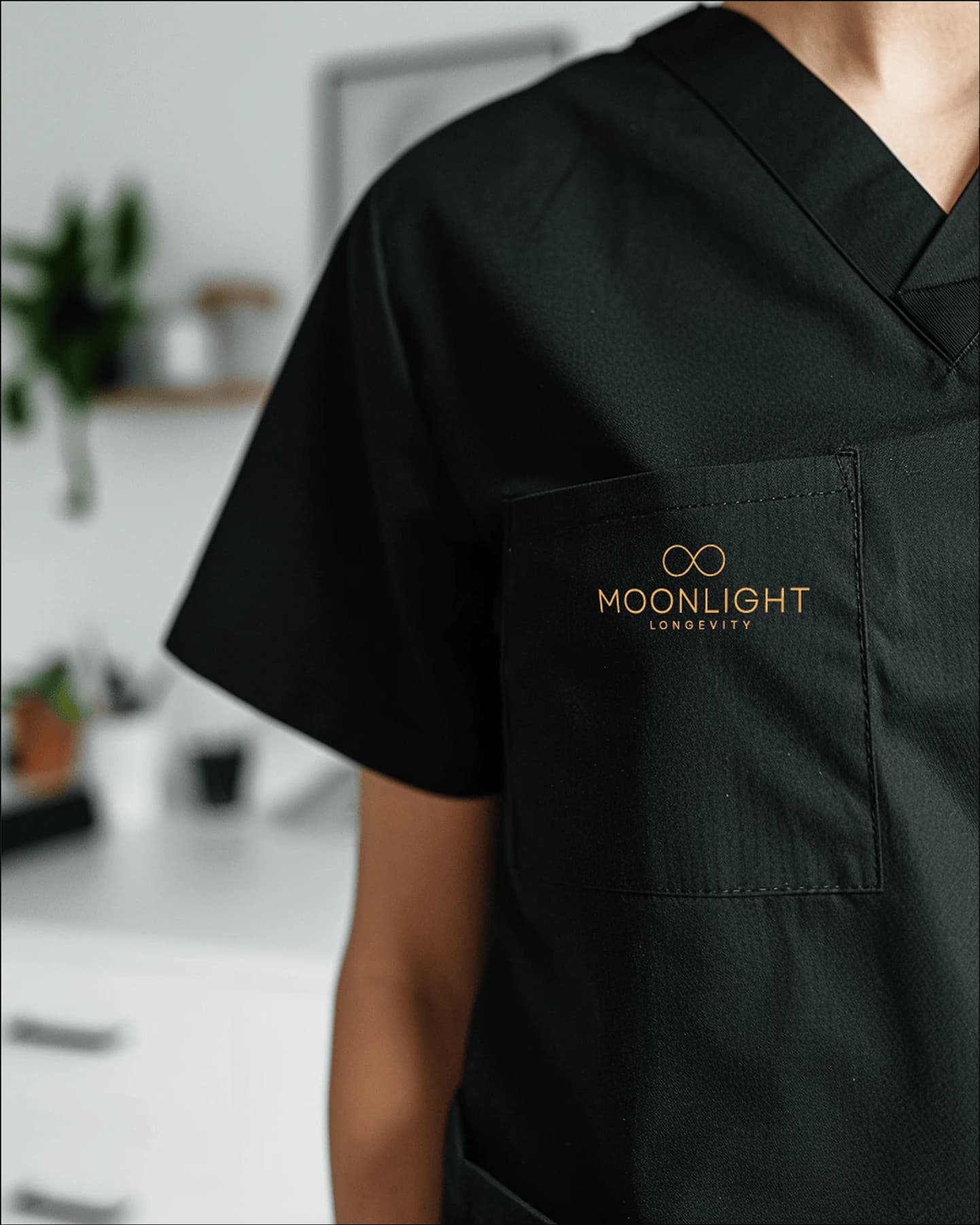 A close-up of the black uniform scrubs with the Moonlight Longevity wordmark embroidered in copper foil on the chest pocket.